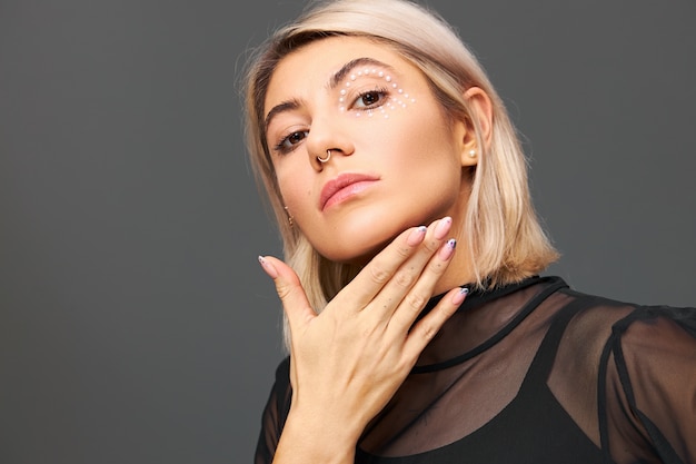 Free photo isolated image of attractive fashionable young female model with facial piercing and stylish make up posing  wearing trendy black transparent blouse. women style, fashion and glamour concept