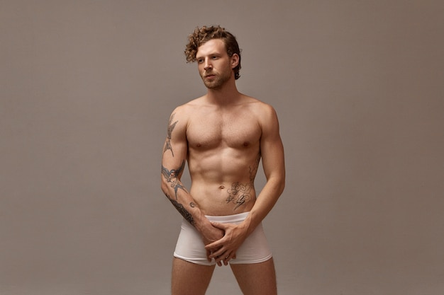 Isolated image of gorgeous handsome man with stubble and curly hairstyle posing naked wearing only white boxer shorts, holding both hands on groin area, having serious facial expression