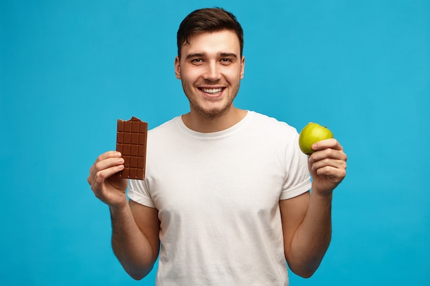 Isolated image of handsome emotional young man keeping strict sugar free diet holding green apple and bar of milk chocolate, having excited expression, going to eat forbidden food as cheat meal