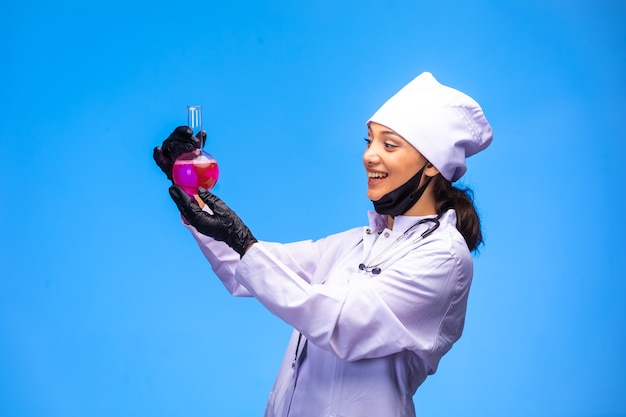 Isolated nurse in hand and face mask shows flask with pink liquid and smiles.