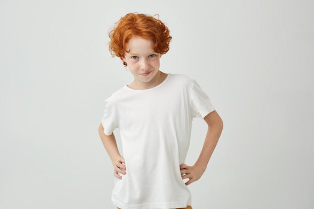 Free photo isolated portrait of funny ginger boy in white t-shirt holding hands on waist