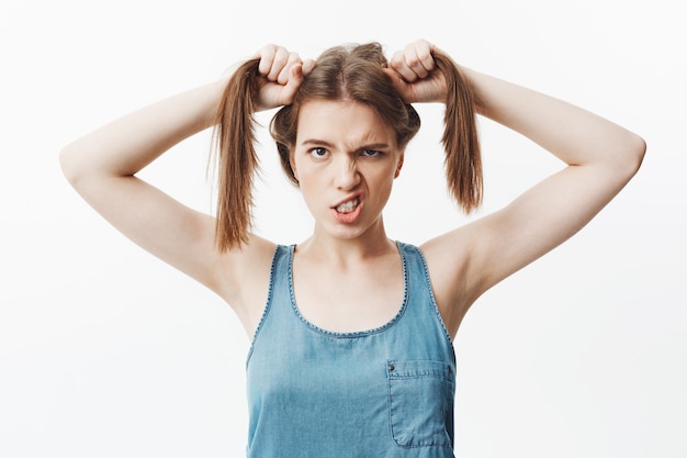 Free photo isolated portrait of funny young caucasian woman with dark hair in bright blue shirt holding hair with hands,  with silly face expression, showing teeth.