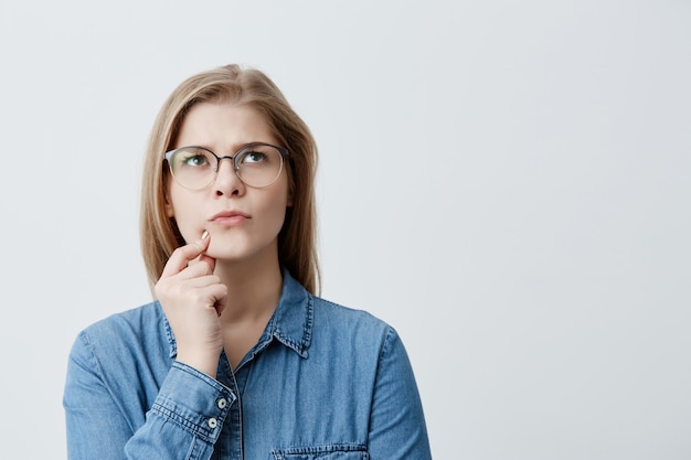 Free photo isolated portrait of stylish young blonde girl in denim shirt and eyeglasses touching her chin and looking sideways with doubtful and sceptical expression, suspecting her boyfriend of lying to her