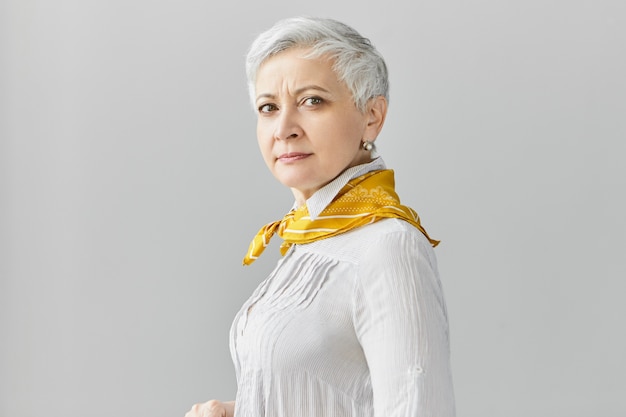 Free photo isolated shot of attractive confident middle aged 60 year old female with short gray hair going to have walk, wearing blouse and silk scarf, posing against blank copyspace wall