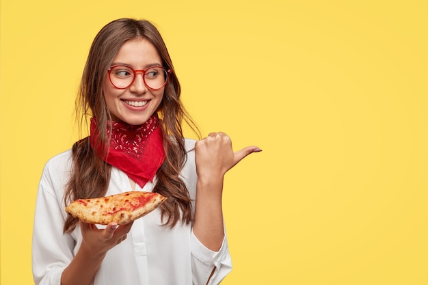 Free photo isolated shot of attractive smiling girl shows direction to pizzeria, eats tasty pizza with cheese and tomatoes, points with thumb at copy space against yellow wall. woman has snack indoor