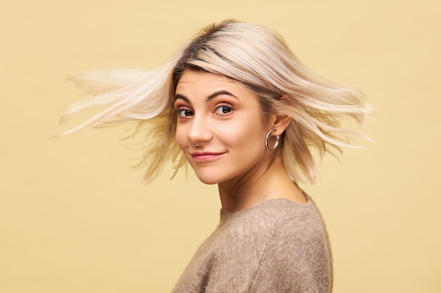 Free photo isolated shot of beautiful amazing young female in stylish oversize sweater turning around with blonde hair flowing in wind