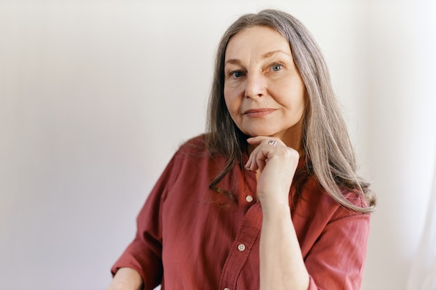 Isolated shot of beautiful thoughtful Caucasian senior female with long gray hair posing at blank wall with copy space for your content, holding hand under chin, having pensive facial expression