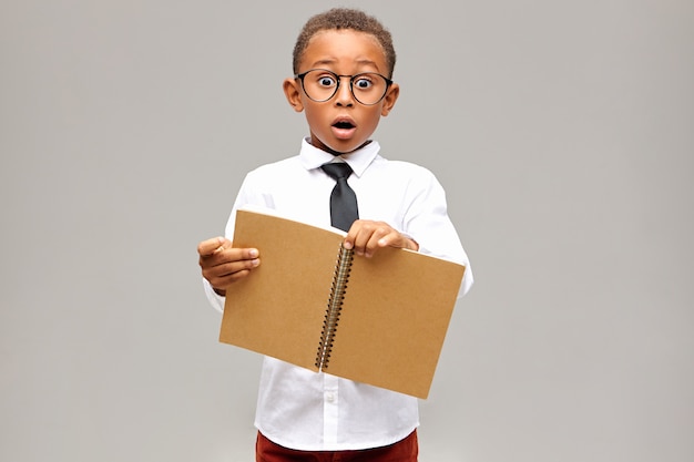 Isolated shot of emotional shocked African pupil wearing white shirt, black tie and eyeglasses having surprised astonished look, keeping mouth widely, holding open blank copybook in his hands