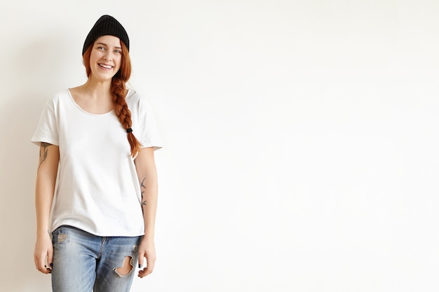 Free photo isolated shot of happy girl wearing her ginger hair in braid, posing at white wall