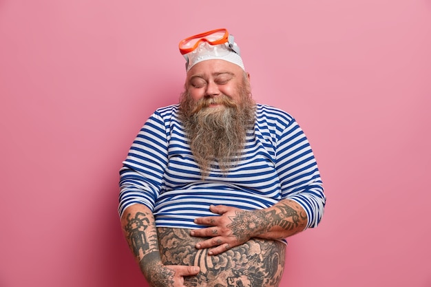 Free Photo isolated shot of plump bearded man holds his fat tattooed belly closes eyes with satisfaction dressed in undersized sailor shirt rubber swimhat and goggles enjoys snorkeling hobby during summer time