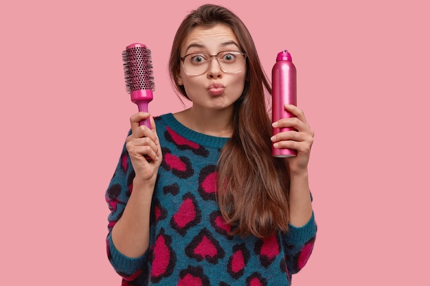 Isolated shot of pretty woman gives mwah, folds lips, has long cared hair, holds comb and hairspray, wears big optical spectacles