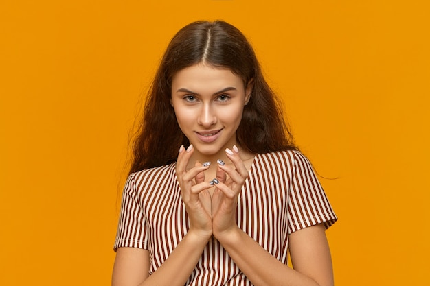 Free Photo isolated shot of smart cunning young female wearing stylish dress with vertical stripes clasping hands