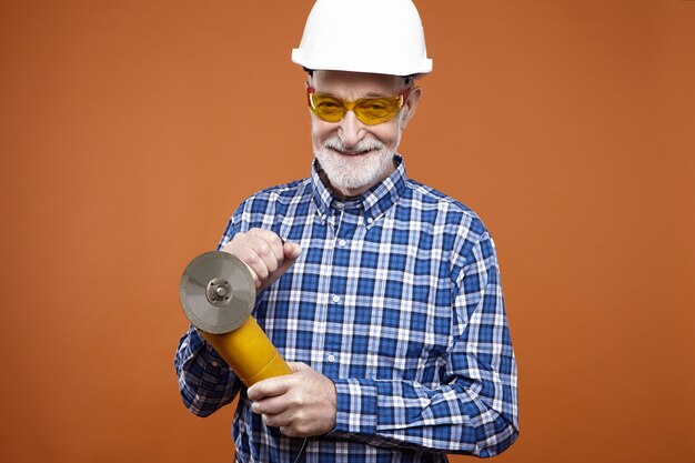 Isolated shot of smiling unshaven aged Caucasian handyman or fitter wearing safety helmet and glasses using angle grinder for cutting and grinding. Heavy duty work, construction and metal concept