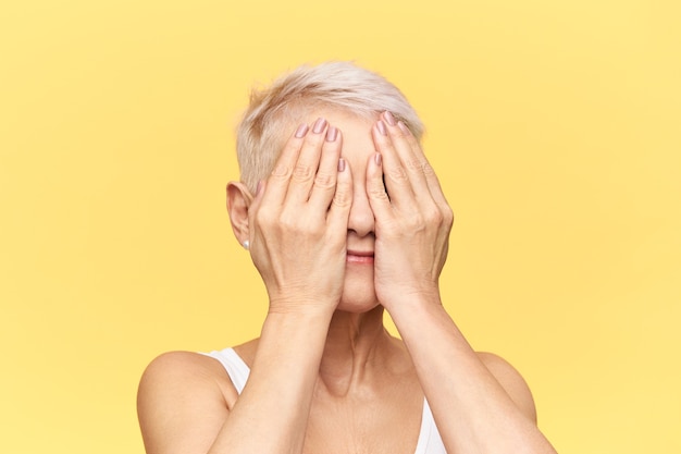 Free photo isolated studio image of unrecognizable grandmother with short blonde hair playing hide and seek with her grandchildren, covering eyes with both hands.