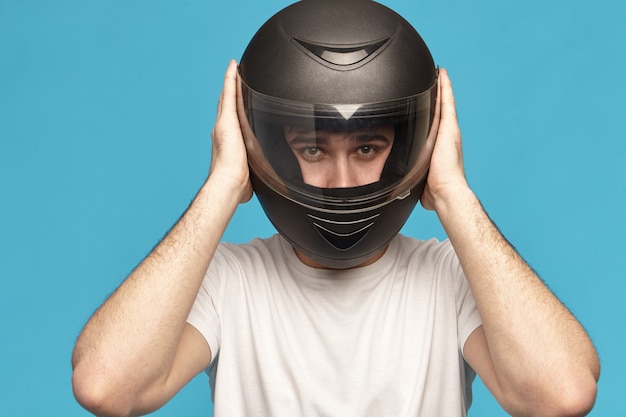 Free photo isolated studio shot of self determined serious young caucasian male biker taking on stylish black motorcycle helmet