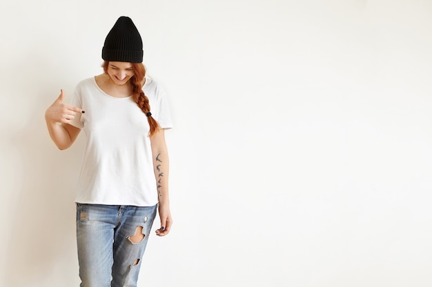 Free Photo isolated studio shot of young female with braid looking down as she pointing at her blank white t-shirt