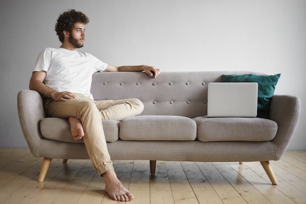 Free Photo isolated view of thoughtful young man with bare feet sitting indoors on sofa with open generic laptop, having small break while working distantly from home. people, job, occupation and technology