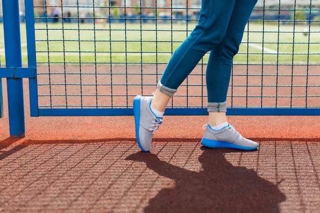 It is a picture of girl`s legs walking near blue fence on the stadium. She wears gray sneakers with a blue line and blue pants.