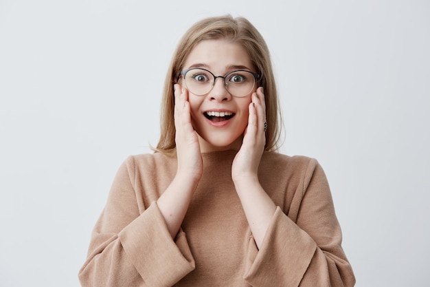 It`s unbelievable! Surprised stunned emotional woman keeps both hands on cheeks, being shocked to see high prices, isolated against blank studio wall. Blonde female can`t believe in shocking news