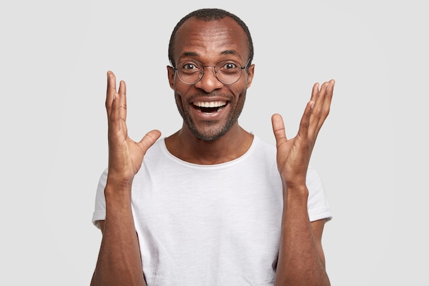 Its amazing and awesome! Joyful dark skinned man raises hands with happiness, has positive expression