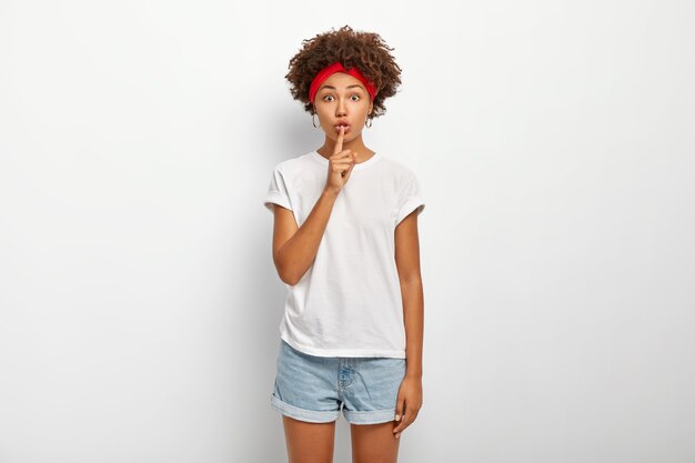 Its better to keep quiet. Intensed bothered woman has Afro hairstyle, explains taboo, makes silence gesture, shushing sign, wears casual outfit, poses against white wall. Keep your voice down