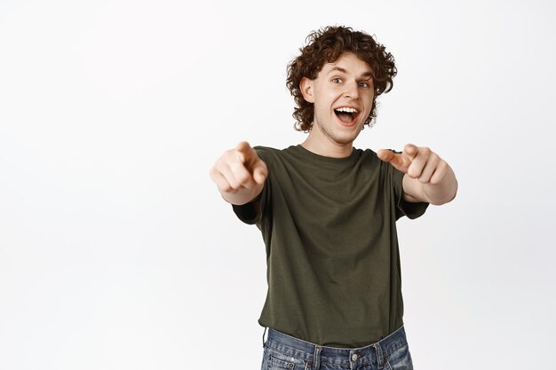 Its you Excited young man pointing fingers at camera shouting ehtusiastic inviting or congratulating people standing over white background