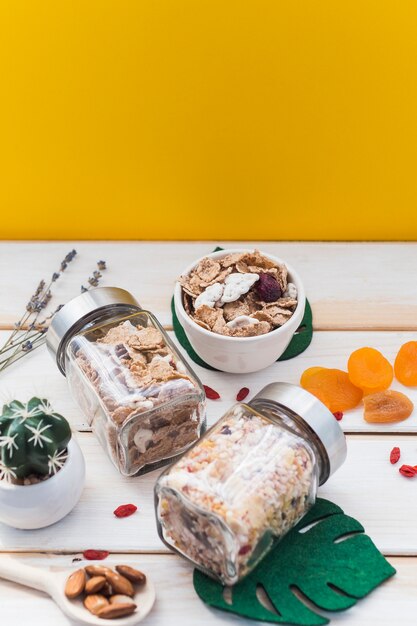 Jar of granola and cornflakes near dry fruits; fake leaf and succulent plant on wooden surface