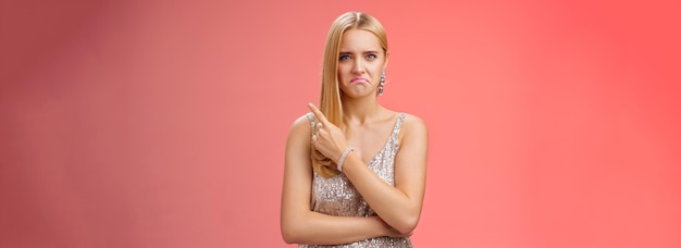 Free Photo jealous displeased angry young revengeful blond exgirlfriend in luxurious silver shiny dress frowning pouting pointing upper right corner displeased pissed standing fed up upset red background