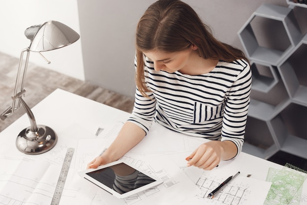 Job, career and business concept. Portrait of young fashionable professional female designer sitting at table, looking in digital tablet monitor, chatting with customer to decide some details.