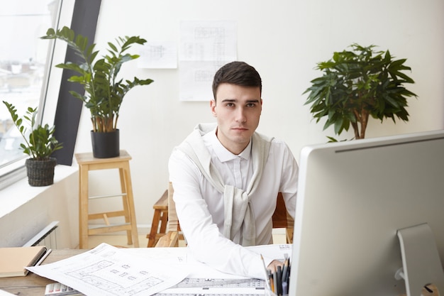 Job, career and success concept. Indoor shot of talented young male architect wearing white shirt and sweater tied around his neck using CAD program on computer while working on construction project