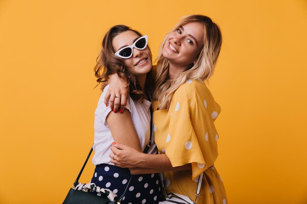 Jocund blonde woman in yellow dress embracing her best friend Inspired darkhaired lady in sunglasses chilling with sister