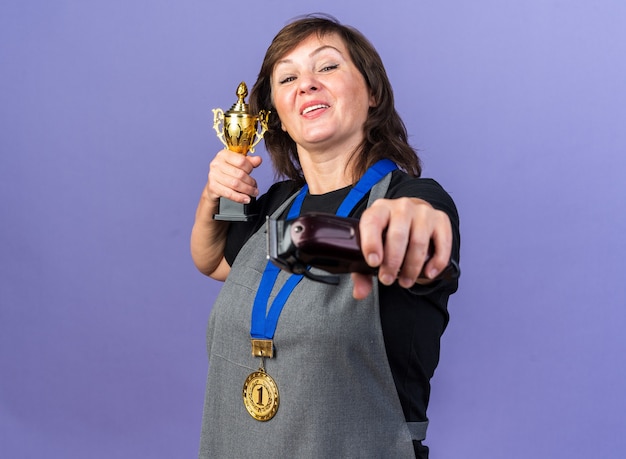 Free Photo joyful adult female barber in uniform with golden medal around neck holding hair clipper and winner cup isolated on purple wall with copy space