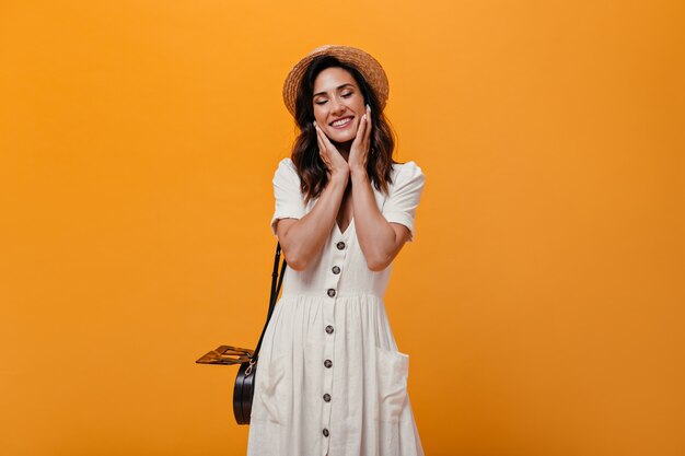 Joyful adult girl in white dress cute smiles on orange background. Pensive woman in small straw hat with black bag posing.