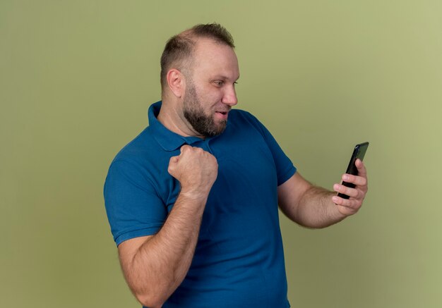 Joyful adult slavic man holding and looking at mobile phone doing yes gesture 