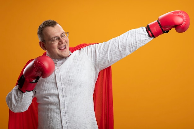 Joyful adult slavic superhero man in red cape wearing glasses and box gloves stretching out hand looking at side keeping another hand in air isolated on orange background