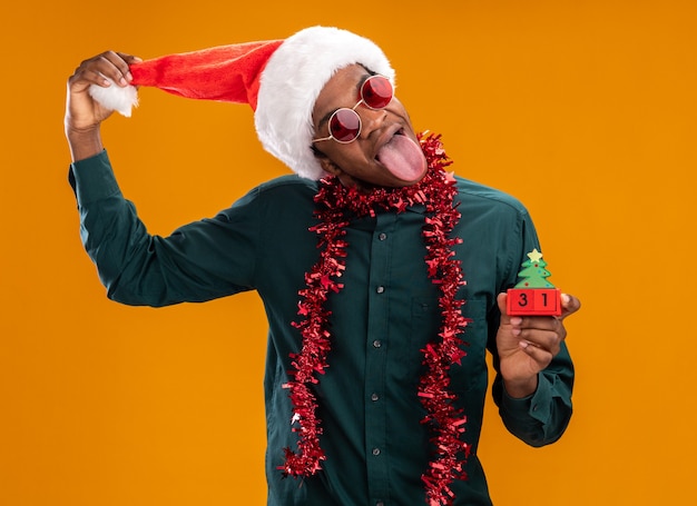 Free Photo joyful african american man in santa hat with garland wearing sunglasses holding toy cubes with new year date sticking out tongue standing over orange background