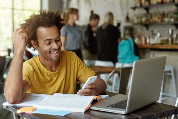 Joyful African American student sitting at wooden table in cafe surrounded with books, exercise books, laptop holding cell phone in hand looking gladly