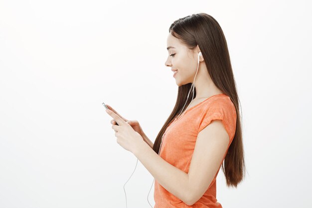 Joyful attractive woman standing in profile over gray background holding smartphone looking at screen with sensual caring smile listening to music in earphones