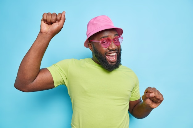 Joyful bearded man dances carefree keeps arms raised up feels happy smiles happily wears pink panama casual green t shirt sunglasses has dark skin isolated over blue wall