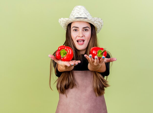 Joyful beautiful gardener girl in uniform wearing gardening hat holding out peppers at camera isolated on olive green background