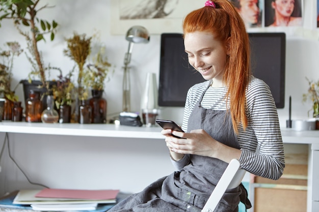 Joyful beautiful young female artist in grey apron relaxing on chair with gadget during small break while working on painting in workshop