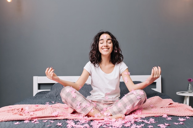 Free Photo joyful beautiful young woman in pajamas with curly brunette hair meditating on bed in pink tinsels. happy model smiling with closed eyes, expressing true emotions, enjoying home comfort