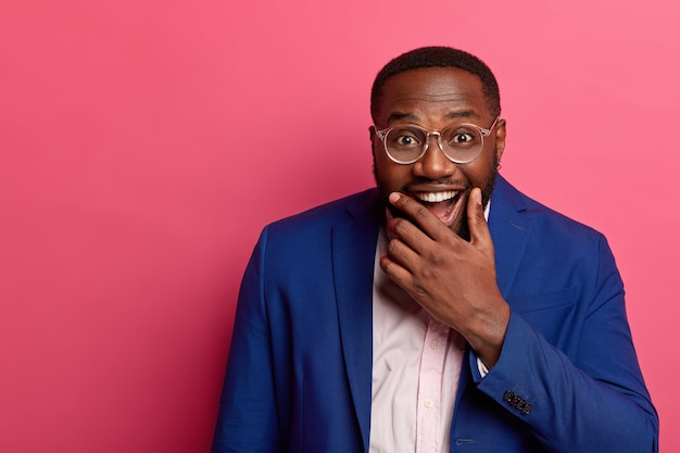 Joyful black man boss holds chin and laughs happily, dressed formally, has pleased funny expression, laughs at joke