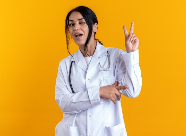 Joyful blinked young female doctor wearing medical robe with stethoscope showing peace gesture isolated on yellow wall