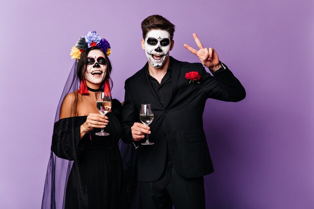 Joyful boyfriend and girlfriend drink champagne and celebrate Halloween in image of Mexican-style bride and groom.