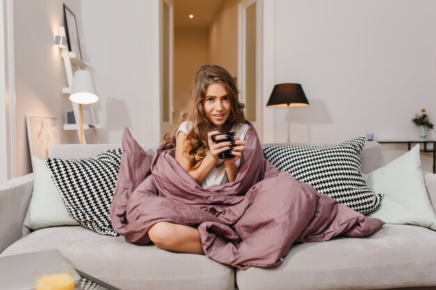 Joyful brown-haired young woman posing with interested face expression, while sitting under blanket