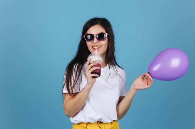 Joyful brunette in black sunglasses looks happy posing with a cocktail and balloon