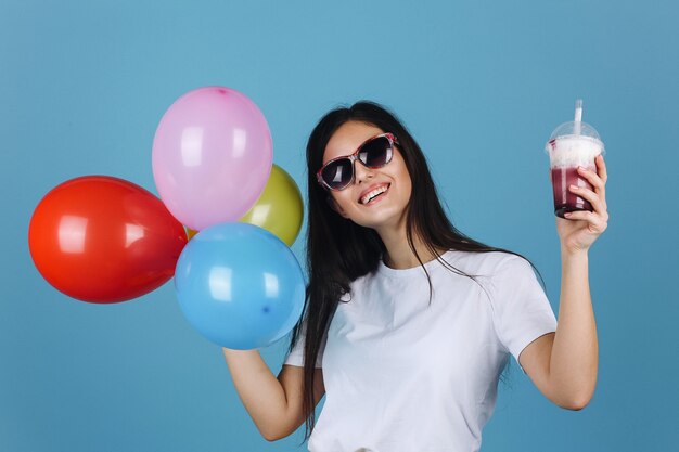 Joyful brunette in black sunglasses looks happy posing with a cocktail and balloons