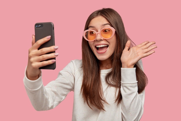 Joyful brunette woman wears sunglasses, makes video call, connected to wireless internet, dressed in white jumper, isolated over pink wall. Hello, friend