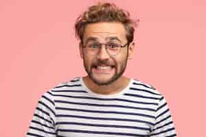 Free photo joyful caucasian male clenches teeth and looks positively, has curly hair, wears spectacles and striped sweater, isolated over pink wall. happy man freelancer rejoices success